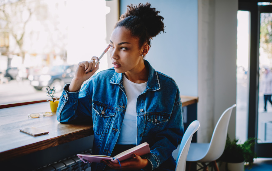 Woman in a denim jacket sits at a cafe window holding a pink notebook and pen while looking thoughtfully to the side.