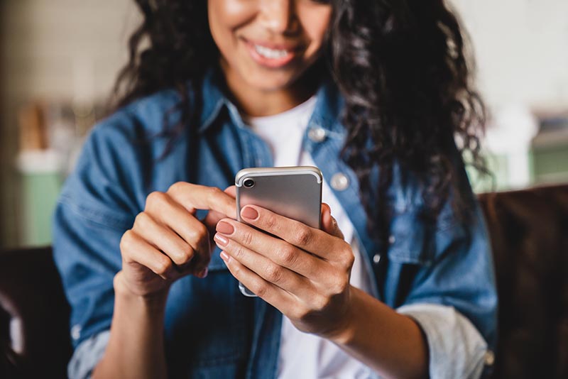 Cropped shot of an african-american young woman using smart phone at home. Smiling african american woman using smartphone at home, messaging or browsing social networks while relaxing on couch Young african-american woman smiling as she looks at the screen on her cell phone.