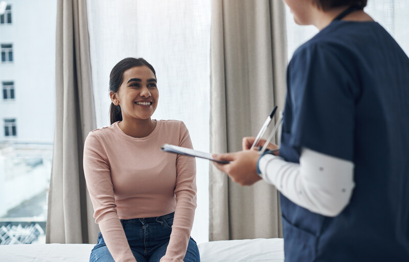 AdobeStock_497158947 (1) A smiling woman sitting on an exam table looks up at a healthcare worker holding a clipboard and pen.