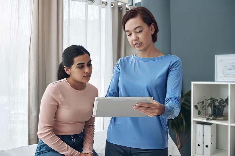 A woman in a blue shirt shows information on a digital tablet to another woman in a light pink shirt indoors.