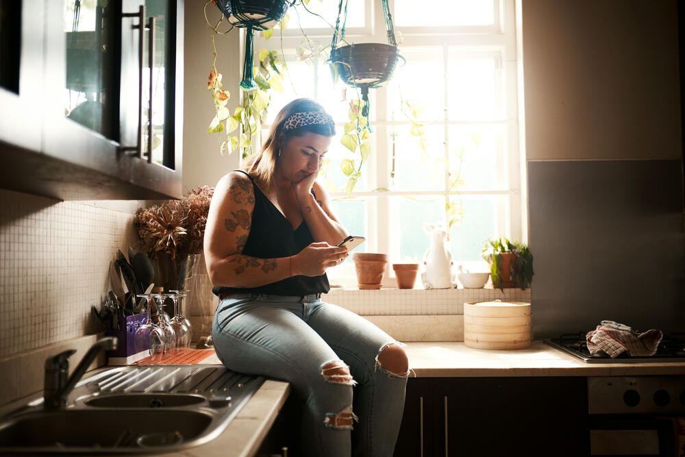 AdobeStock_522158168 (1) A woman with tattoos sits on a kitchen counter near a bright window while looking at her smartphone.